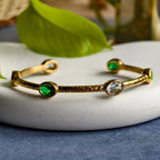 Gold cuff bracelet with green and clear oval gemstones on white plate with green leaf background
