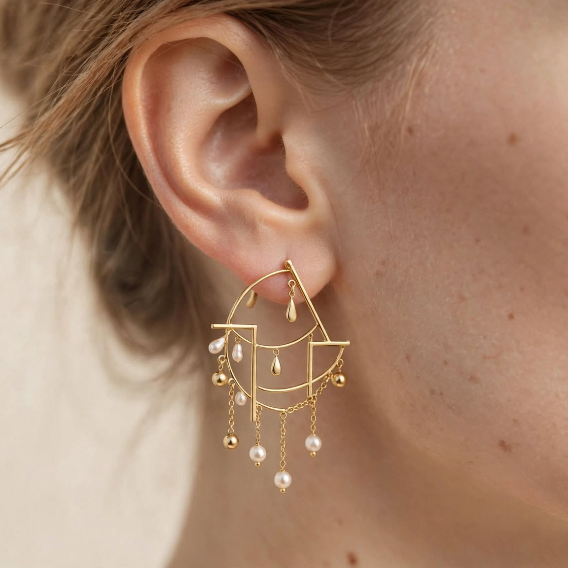 A close-up of a model's ear wearing a gold geometric dangle earring with small pearl-like beads, against a soft beige background.