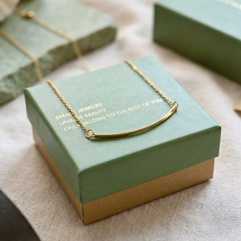 Pretty Shimmering Things Global Sunrise Curve gold bar necklace resting on a green gift box with minimalist design.